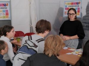 Une animatrice souriante présente le Pique-nique linguistique à un groupe de jeunes visiteurs attablés.